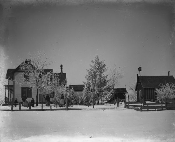 Members of the Krueger family standing in the fenced-in yard of Alexander Krueger's home. The home and several farm buildings are behind the family. Fresh snow covers the trees making them white.