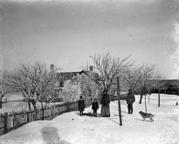 Jennie, Edgar, Mary, and Alexander Krueger standing in an old orchard on their property during winter. Between Edgar and Jennie is a sled, and a dog is standing on the right looking at them. On the left is a fence with the farmhouse in the background.