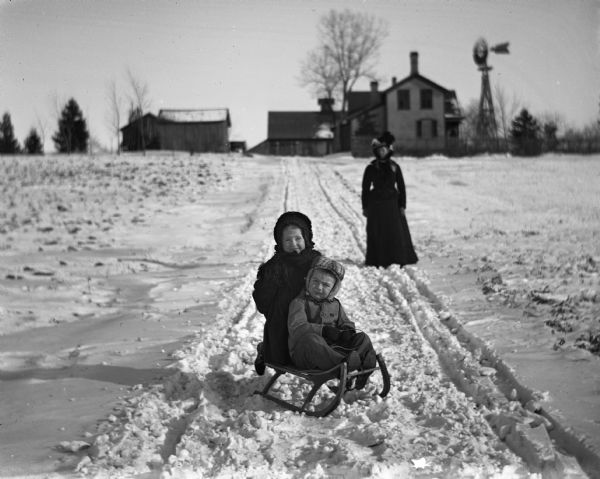 Winter scene with Jennie and Edgar Krueger posing on a sled in the snow on the west driveway of the family farm. Edgar sits in front of the sled, while Jennie sits behind him. Florentina Krueger stands further up the driveway. The house, farm buildings, and a windmill are up a slight incline at the end of the driveway.