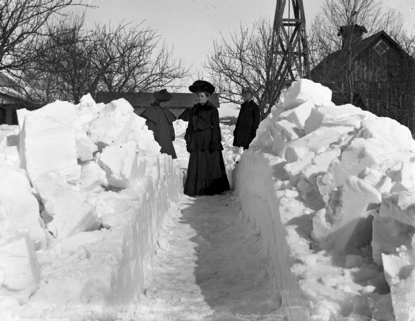 Winter scene with Jennie, Flora and Edgar Krueger standing at the end of a path shoveled in extremely deep snow. Farm buildings and the base of what may be a windmill are in the background.
