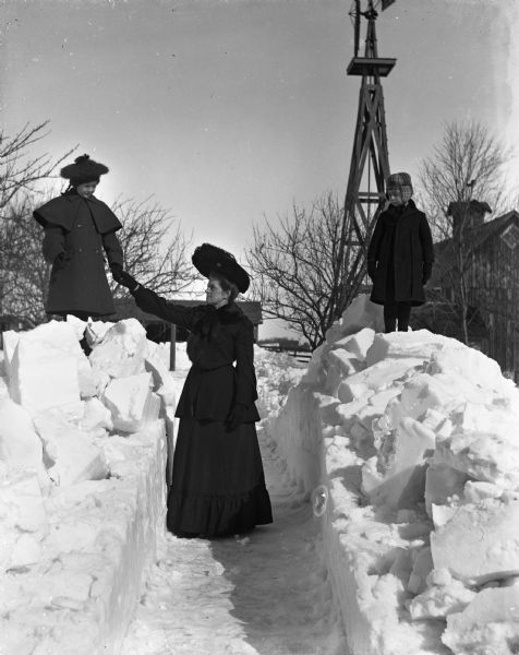 Winter scene with Jennie and Edgar Krueger standing on top of blocks of snow on either side of a shoveled path through deep snow in front of the Krueger house. Florentina is standing between the two on the shoveled out section of the path. She is holding Jennie's hand.