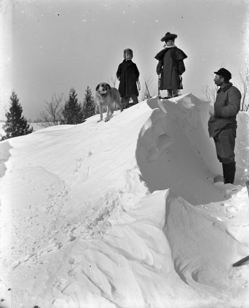 Winter scene with Jennie and Edgar Krueger standing on top of a large snow drift with one of their dogs. They each appear to be holding the ropes of sleds. Alexander Krueger is standing in profile below them on the right.