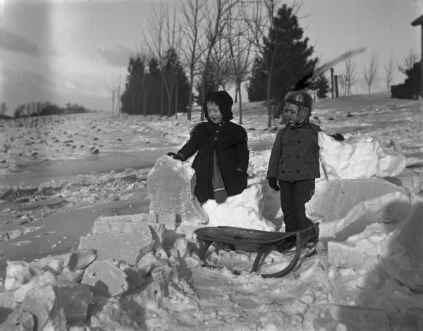 Winter scene with Jennie and Edgar Krueger (twins) playing with large blocky pieces of snow in a field. A sled is on the ground in front of them.