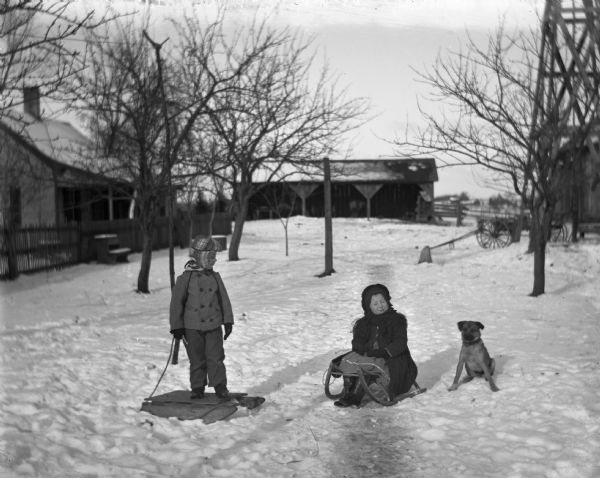 Winter scene with Jennie and Edgar Krueger in the yard with their sleds. Jennie is on the right sitting on her sled, and a dog is sitting in the snow next to her. On the left Edgar is standing on top of his sled. In the background are trees and farm buildings.