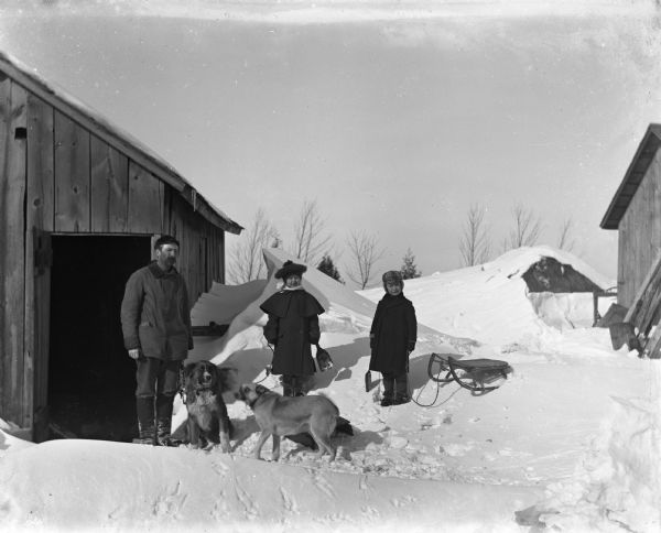 Winter scene with Jennie and Edgar Krueger (twins) standing upon snow that has drifted against the woodshed. Each is holding a shovel and a rope that is attached to their sleds. Alexander Krueger is standing next to them with two of his dogs.