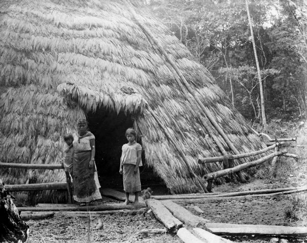 A women and her two children standing in front of the doorway to a traditional home of the indigenous people of Costa Rica. Several logs act as a sidewalk to the front door.