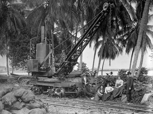 Two men and three women posing next to a crane on a railroad track along the coast of Costa Rica. Coconut palms are behind them along the Caribbean Sea. The city of Limon is in the background along the far shoreline.
