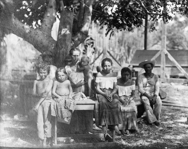 Several Costa Rican villagers posing together next to a tree on a table and a bench. A woman stands behind the table and supports an infant standing on it. Several children stand sit along its edge. A man and woman sit on a bench on the right. Village buildings stand along the edge of the forest in the background.