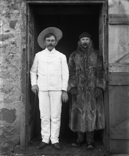 William Wendorf and Alexander Krueger standing together in an open barn doorway. William is dressed in his white suit from Costa Rica, with a straw hat. He is also holding a cane. Alexander is dressed in his winter clothing of a long fur coat, hat, gloves, and boots.