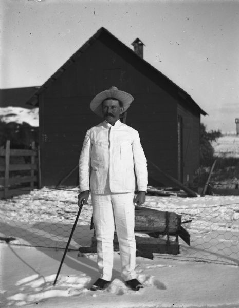 William Wendorf standing outside in the snow near a fence and a shed.  He is wearing his white suit from Costa Rica and a straw hat, and is holding a cane in his right hand.