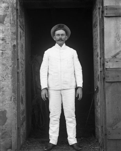 William Wendorf standing in a barn doorway wearing a white suit from Costa Rica. His cane is resting against the doorway on the right.