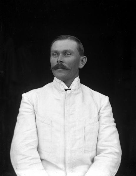 Waist-up portrait in front of a dark background of William Wendorf wearing his white suit from Costa Rica.