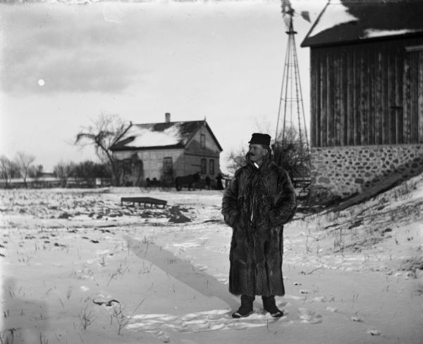 William Wendorf standing in the snow in front of barn and windmill wearing a long fur coat. A horse pulling a buggy is standing with two individuals near the half timber house in the background.