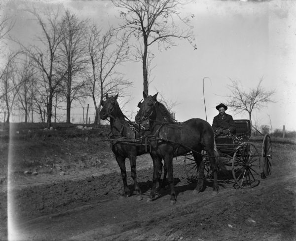 August Kressin driving a buggy that is pulled by a team of horses along a dirt road. Trees and a fence are in the background.