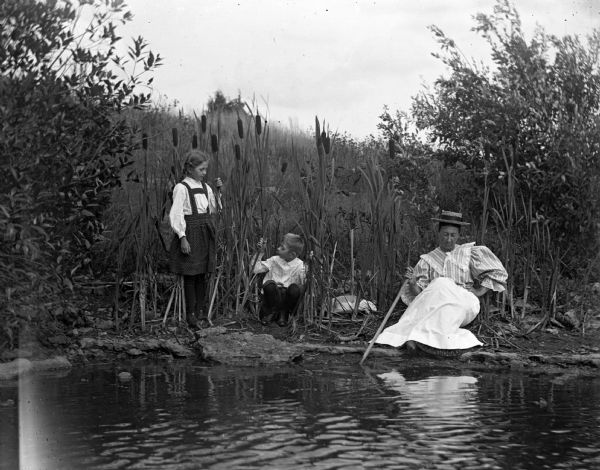 View over water towards Jennie and Edgar Krueger playing amongst the cattails at water's edge while their grandmother, Mary Krueger, is sitting along the edge of the quarry pond playing with a stick in the water. Jennie's hat is sitting on the ground among the cattails.