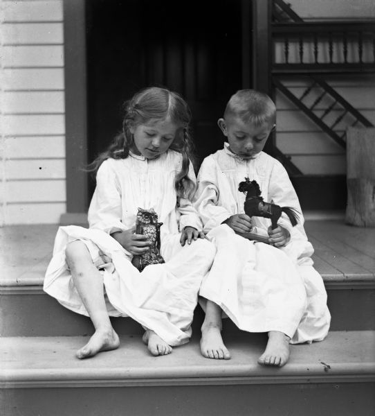Jennie and Edgar Krueger sitting on the porch steps in their night gowns. Both are looking down at a toy they are holding in their hands, Jennie an owl and Edgar a horse.