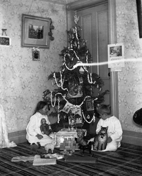 Jennie and Edgar Krueger sitting on the floor in their nightgowns next to the family Christmas tree with their presents. Each of them is holding a stuffed toy.