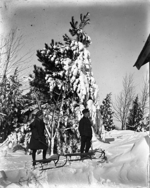 Winter scene with Jennie and Edgar standing on a snowdrift with their sleds. A snow-covered pine tree is behind them. A small building is behind the pine tree, and the roof of another building is on the right.