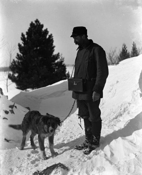 Winter scene with Alexander Krueger standing on a snowdrift looking down at his dog standing beside him. Alexander has a camera case around his neck.