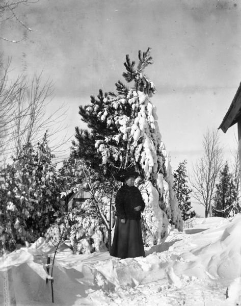 Outdoor portrait of Sarah Krueger standing next to a snow-covered pine tree. There is a small farm building behind the pine tree.