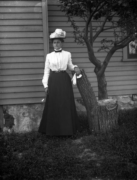 Full-length outdoor portrait of Florentina Krueger standing while resting her right arm on a log stump shaped into a chair. She is holding a pair of white gloves in her right hand. The side of the house is behind her.