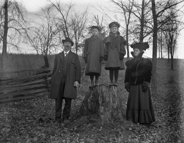 Outdoor portrait of the Alexander Kruger family standing near a tree stump with a split-rail fence behind them. Alexander and Florentina Kruger are standing on either side of the stump, and the twins, Jennie and Edgar, are standing on top of the stump.