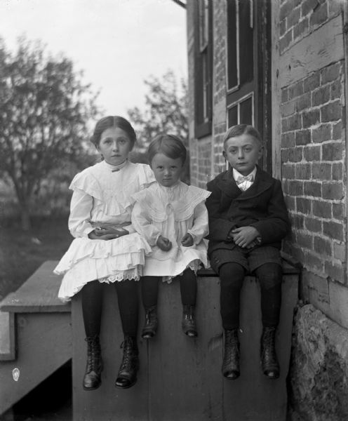 Jennie and Edgar Krueger sitting and hanging their feet off the edge of the side of wooden front steps leading up to a brick home, with Ruth Kressin sitting between them.