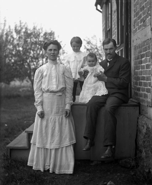 Outdoor portrait of the August Kressin family posing on the side of the front steps of a house. August Kressin sits on the top step holding Alice Kressin, while Ruth Kressin is standing behind him. August's wife, Martha Wendorf Kressin, is standing in front of them. The brick house is half-timbered.