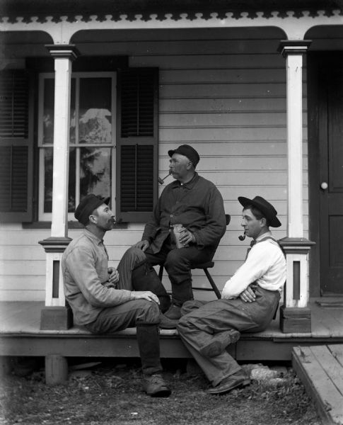 Alexander Krueger, August Krueger, and Henry Bigalk sitting on the front porch smoking pipes. August is sitting on a chair holding the bag of tobacco, while the other two men are sitting on the porch floor leaning against support posts.