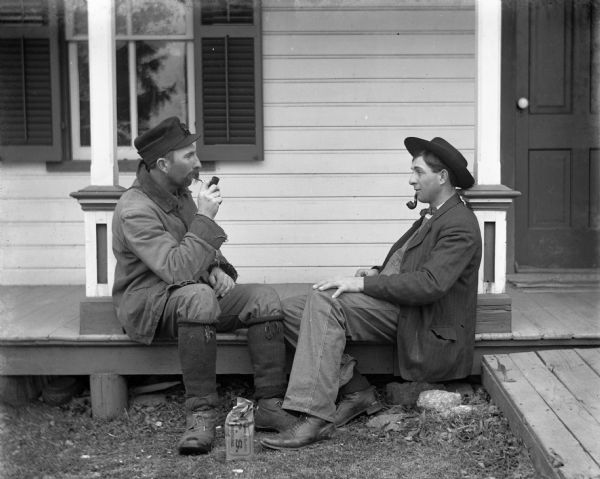 Alexander Krueger and Henry Bigalk sitting on the floor of the front porch smoking pipes. The bag of tobacco is sitting on the ground in front of them.