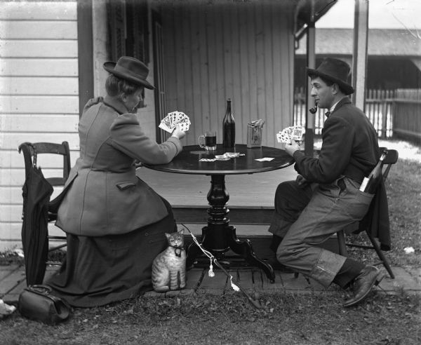 Sarah Krueger and Henry Bigalk playing cards at a table set up outdoors next to the porch. Henry is smoking a pipe and has a large bottle of alcohol sticking out of the back pocket of his overalls, which he has on over his suit pants. On the table is a bottle, a mug, and a bag of tobacco. A stuffed animal cat is leaned up against the side of Sarah's dress, and August Kruger's long pipe is resting on the bottom leg of the table.