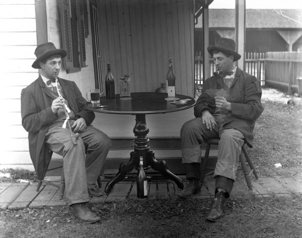 Double exposure image of Henry Bigalk playing cards with himself at a table placed outside next to the porch. Alcohol bottles, cards, a mug, and a bag of tobacco are sitting on the table.