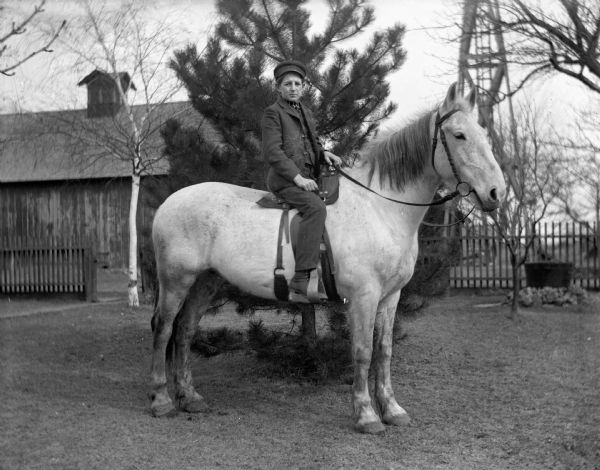 Sam Goetsch riding a horse on a farm. In the background is a barn and what may be the support structure for a windmill.