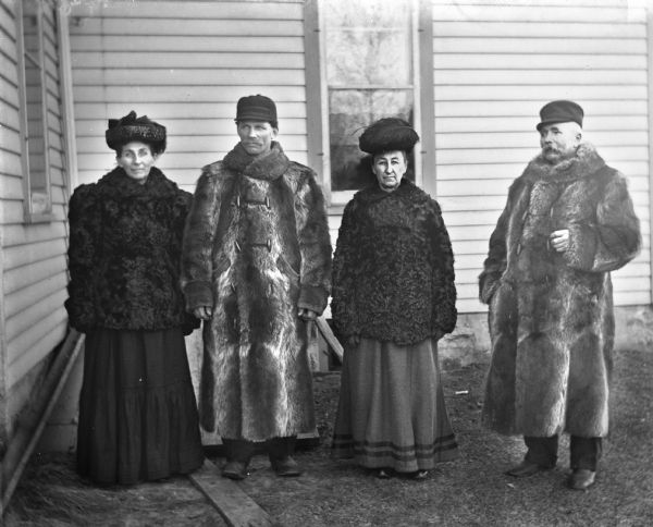 Group portrait of Liddie Blatter, Gottlieb Blatter, Mary Krueger, and August Krueger standing in a yard next to a house wearing fur coats.