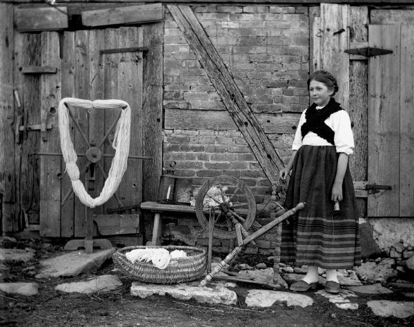 Jennie Krueger standing on stones next to a spinning wheel and yarn winder placed outside a barn. She is wearing her great-grandmother's traditional Pomeranian attire and her wooden soled shoes. A handmade basket made by her great-grandfather is sitting next to the spinning wheel filled with yarn.
