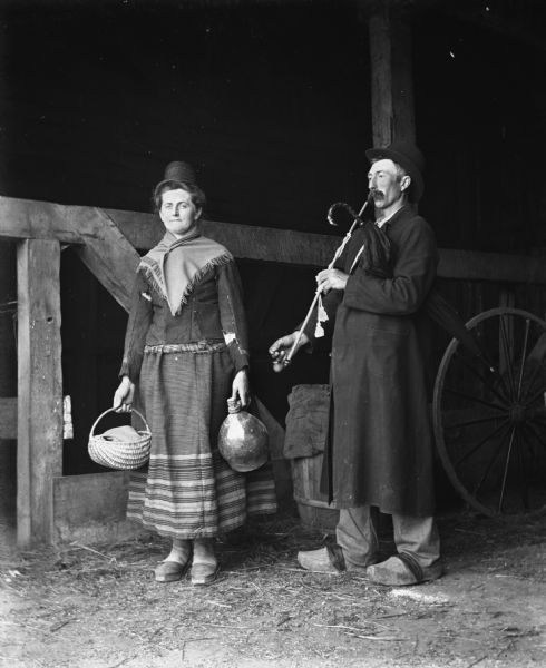 Florentina and Alexander Krueger standing in a barn wearing traditional Pomeranian attire. Alexander is smoking his fathers pipe while holding an umbrella under his arm, while Florentina is holding a jug and a handmade basket made by William Krueger. They are both wearing traditional wooden soled shoes.