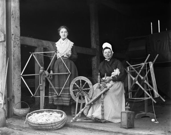 Tille Faulkman, standing, and Sarah Krueger, are sitting behind a spinning wheel and a yarn winder in the open doorway of a barn. Both women are wearing traditional Pomeranian attire.
