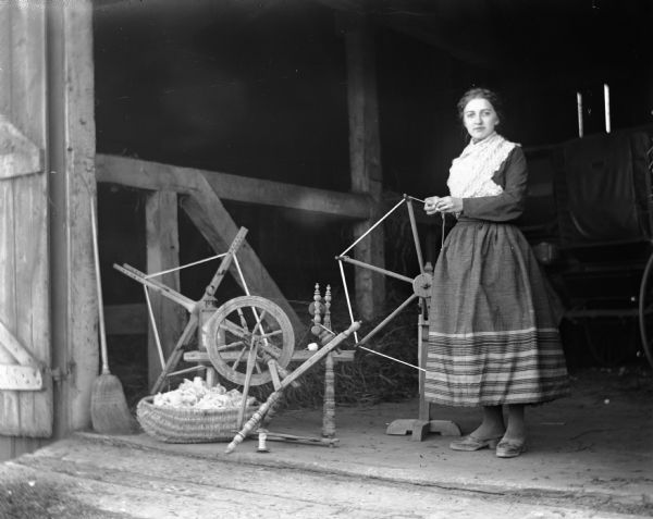 Tille Faulkman standing in a barn with a spinning wheel and two yarn winders. She is dressed in traditional Pomeranian attire. A handmade basket filled with yarn is sitting on the ground next to the spinning wheel.