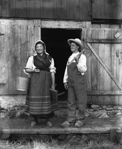 Sarah and Florentina Krueger standing in front of a barn in costume.  Sarah is dressed in traditional Pomeranian attire and is holding a bucket and milking stool. Florentina is dressed in mens overalls and hat with a pitchfork propped over her left shoulder.