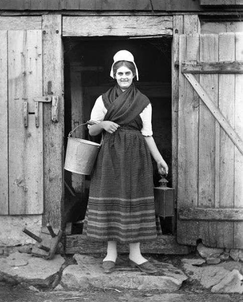 Flora Fels Weege standing in a barn doorway carrying a bucket and a lantern while dressed in traditional Pomeranian attire.