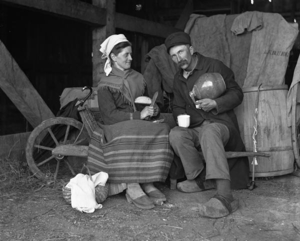 Alexander and Florentina Krueger are sitting in a barn wearing traditional Pomeranian attire, including the wooden shoes. Alexander is pouring himself something to drink from a large jug, while Florentina is cutting a slice of bread.