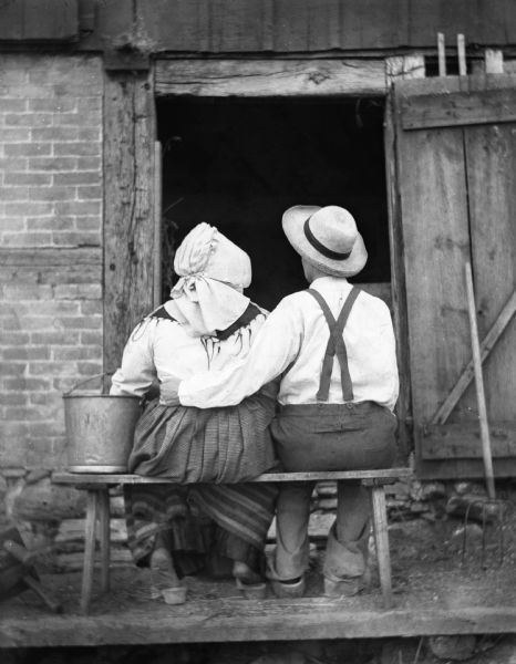 Sarah and Florentina Kruger are sitting on a bench in front of a barn doorway. Florentina is dressed in men's overalls and has her arm around Sarah, who is wearing traditional Pomeranian attire. Both women have their backs to the camera.
