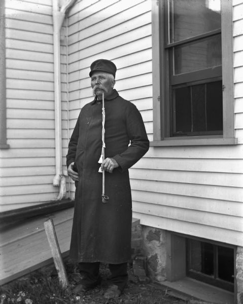 August Krueger standing next to a house wearing a long black coat smoking his pipe.