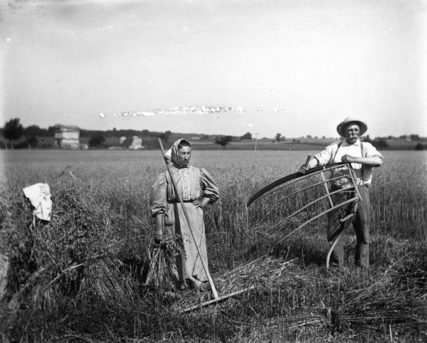 The photographer posing his parents, Mr. and Mrs. August Krueger, cradling and raking grain in a field. She is wearing a dress, and a scarf over her head. He is wearing a hat and trousers with suspenders.