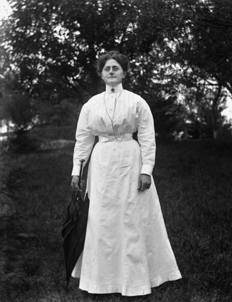 Outdoor portrait of Florentina Krueger standing and wearing a white dress with a black umbrella resting at her side.