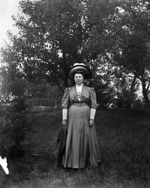 Outdoor portrait of Sarah Krueger standing in a dress and wearing a hat, with an umbrella resting at her side.