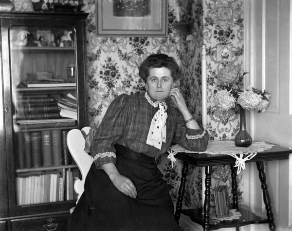 Indoor portrait of Florentina Krueger sitting in a chair, leaning against a side table. A vase with flowers is sitting atop a doily on the side table. A bookcase is behind her.