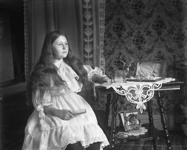 Indoor portrait of Jennie Krueger sitting, with her hair down. She is holding a comb in her right hand and resting her left arm on an end table that is holding a mirror. A box for the mirror and comb is standing open on top of the end table next to a statue of a buck.