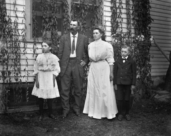 Outdoor group portrait of the Alexander Kruger family. Alexander and Florentina are standing in between their two children, Jennie and Edgar, next to a house with ivy climbing the windows and the siding.