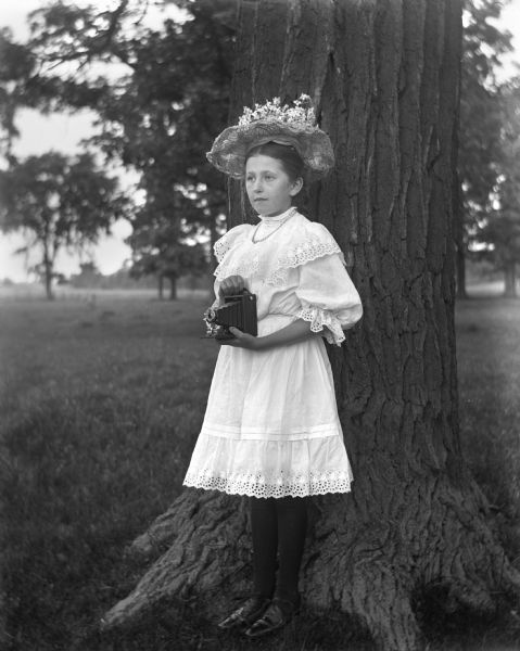 Jennie Krueger standing at the base of a tree, wearing an elaborate hat and holding her fathers' camera.